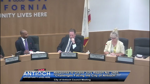 a black man in a suit looking at a white man in a suit with a white woman on his other side. name plates show the black man is lamar a hernandez thorpe mayor, the white man is mike barbanica council member and the woman is lori ogorchok council member. text: antioch opportunity lives here responses to grand jury report number 2405, "challenges facing the city of antioch." city of antioch council meeting