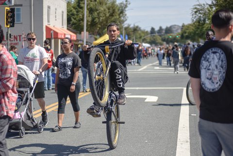 a young man doing a wheelie on a bicycle on a city street with other people walking around