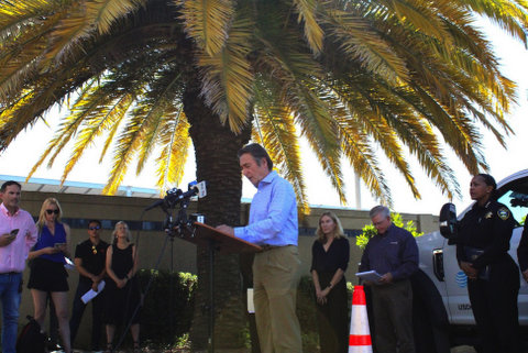 a white man standing in front of microphones under a short palm tree giving a press conference with other people standing around him, including a black woman in police uniform