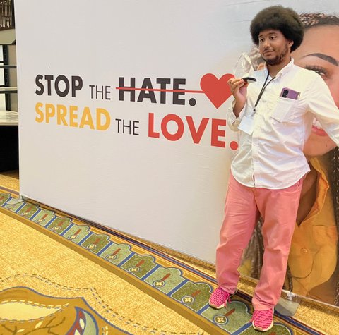 a young black man with an afro wearing a white button down shirt, salmon colored pants and pink shoes, holding a clear plaque and standing in front of a backdrop that says stop the hate spread the love