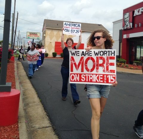 young woman walking with sign that reads "we are worth more on strike for 15" and another woman holding a sign above her head that reads "america can't survive on $7.25." other people are walking with signs as well. they are passing a KFC