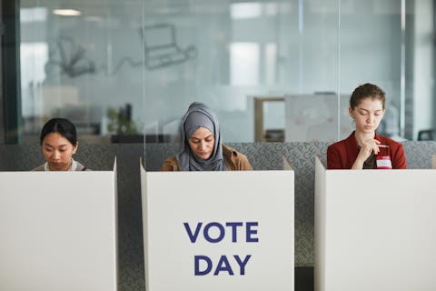 three young women at voting booths. one is east asian, one is wearing a hijab and one is white. the middle booth says vote day