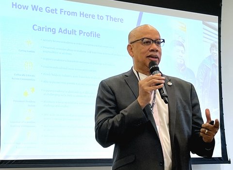 a bald black man wearing a suit jacket and glasses holding a microphone in front of a screen with text that reads how we get from here to there caring adult profile