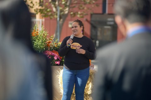 latina woman holding a microphone and wearing a long sleeved black shirt with an illustration of a burger with the words michocan burgers between the buns