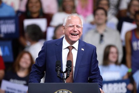 Tim Walz, an older white man who is balding with white hair. He is smiling and wearing a blue suit standing at a lectern that is barely shown with an out of focus crowd behind him