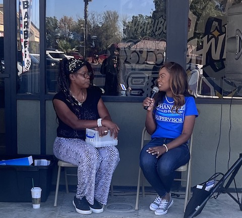 two black women sitting side by side in folding chairs, facing each other. one is holding a microphone and wearing a blue t shirt that says shanelle scales preston supervisor