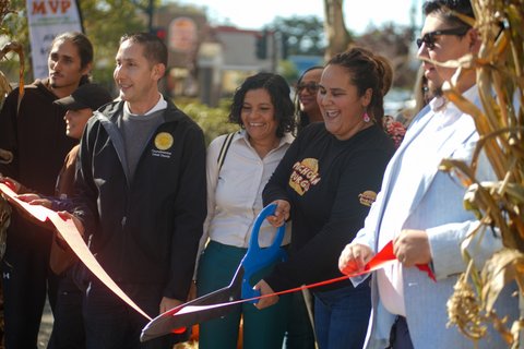 a latina woman cutting a ribbon with large novelty scissors next to another latina and a latino man among a crowd