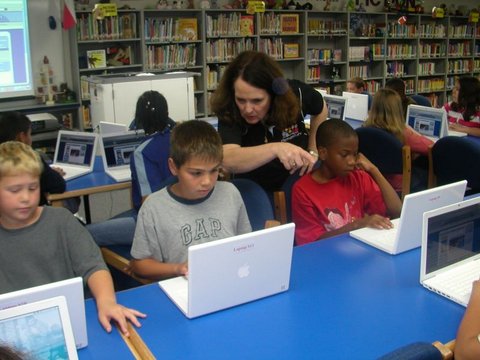 a middle aged white woman standing over two white boys and a black boy using apple laptops with other kids on computers in the background in a school library