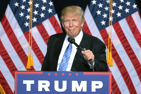 donald trump at a lectern with a trump sign in front of american flags