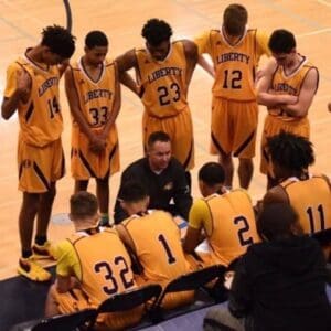a high school boys basketball team on the sideline with a row of five players standing and a row of four players sitting with their coach kneeling between them