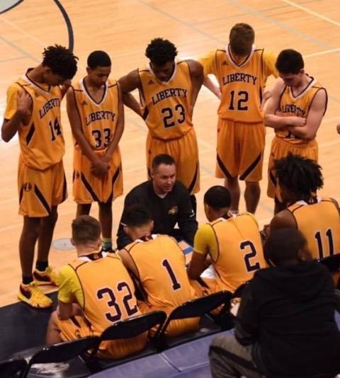 a high school boys basketball team on the sideline with a row of five players standing and a row of four players sitting with their coach kneeling between them