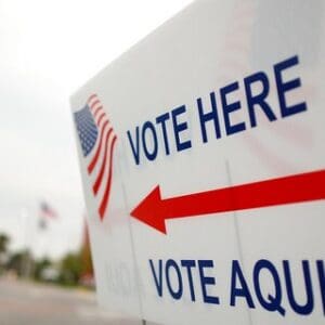 sign that reads vote here vote aqui with red arrow and US flag illustration