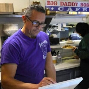 man in purple T shirt and glasses at a tablet register with a restaurant kitchen and sign that says big daddy ross's cafe behind him