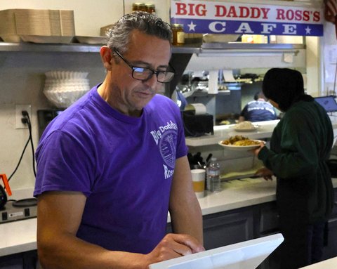 man in purple T shirt and glasses at a tablet register with a restaurant kitchen and sign that says big daddy ross's cafe behind him