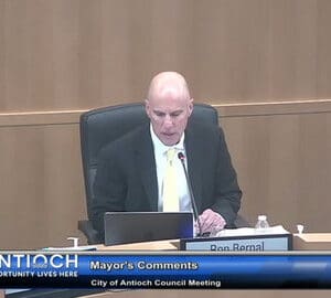 bald man in a suit sitting in government meeting. in front of him are a laptop, which he is looking at, a microphone and a nameplate that says ron bernal. on screen text reads antioch opportunity lives here mayor's comments city of antioch council meeting