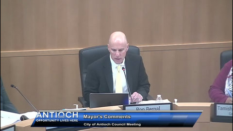 bald man in a suit sitting in government meeting. in front of him are a laptop, which he is looking at, a microphone and a nameplate that says ron bernal. on screen text reads antioch opportunity lives here mayor's comments city of antioch council meeting