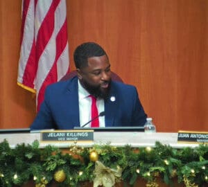 black man in a suit with nameplate that reads jelani killings vice mayor. a US flag is partially visible behind him and christmas greenery and ornaments are on a strand in front of him
