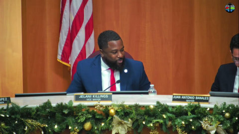 black man in a suit with nameplate that reads jelani killings vice mayor. a US flag is partially visible behind him and christmas greenery and ornaments are on a strand in front of him