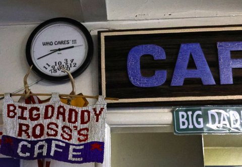 on a wall are a black clock with white face that says who cares!!! in the middle with the numbers scattered at the bottom; a beaded sign that says big daddy ross's cafe; and two other signs, the visible portions of which say CAF and big dad