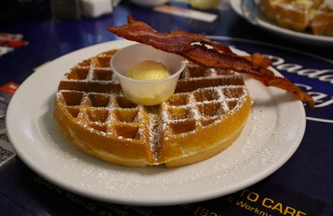 a large round waffle on a white plate sprinkled with powdered sugar. butter is in a plastic cup in the center of the waffle and a strip of bacon is leaning against the cup and waffle