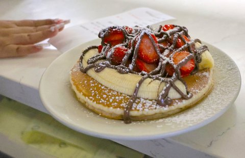 pancakes on white plate topped with bananas, strawberries, chocolate syrup and powdered sugar