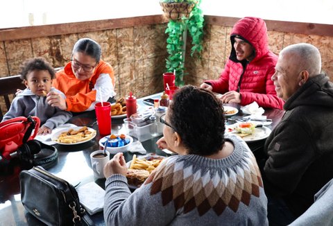 four adults and a child sitting around a table of food. a woman in an orange and white san francisco giants jacket is feeding the kid a french fry