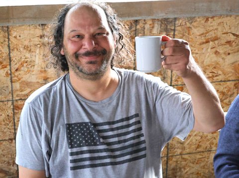 a white man wearing a gray T shirt with colorless USA flag design smiles while holding up a white coffee mug