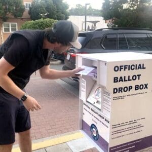 a white man placing an envelope into an official ballot drop box