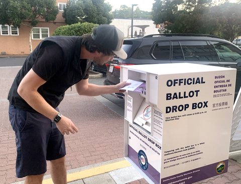 a white man placing an envelope into an official ballot drop box