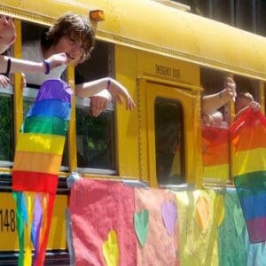 I Thought Pride Didn’t Include Me. I Was Wrong close up of a yellow school bus with a makeshift rainbow flag duct taped to the side. the flag is decorated with hearts and appears to have been written on. a boy is leaning out of a window and several other people are holding their arms out, waving or holding rainbow flags