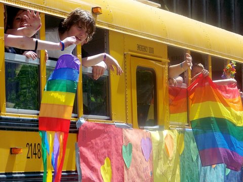 close up of a yellow school bus with a makeshift rainbow flag duct taped to the side. the flag is decorated with hearts and appears to have been written on. a boy is leaning out of a window and several other people are holding their arms out, waving or holding rainbow flags