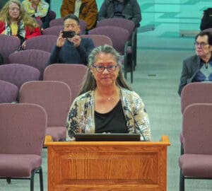 an older japanese woman standing at a lectern with other people seated behind her