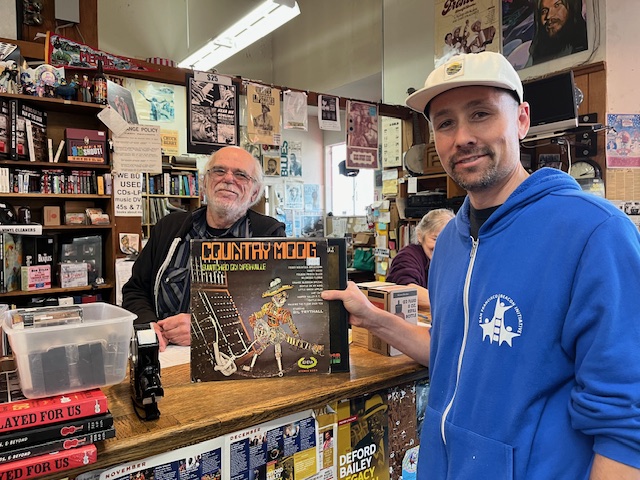 a white man holds a vinyl album titled country moog on the counter of a record store behind which is a balding white man with white hair and beard and glasses