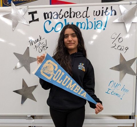 a latina teen holding a columbia pennant flag in front of a white board that is decorated with silver stars and has the following written on it i matched with columbia, thank you QB, CO 2029, NYC here I come