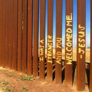 fence on US mexico border with the following painted across multiple pillars: i was a stranger and you welcomed me — jesus