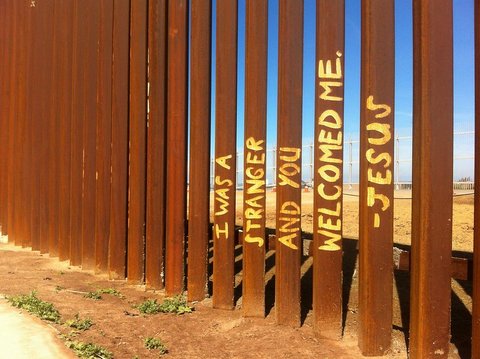 fence on US mexico border with the following painted across multiple pillars: i was a stranger and you welcomed me — jesus