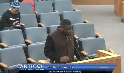 black man standing at a lectern with onscreen text that reads antioch opportunity lives here public comments city of antioch council meeting. behind him a black woman is sitting among mostly empty seats