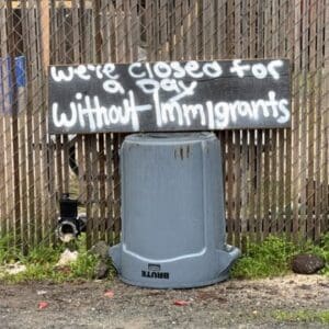 a wooden board with we'e closed for a day without immigrants spray painted on it. the sign is perched on an overturned gray trash can in front of a fence