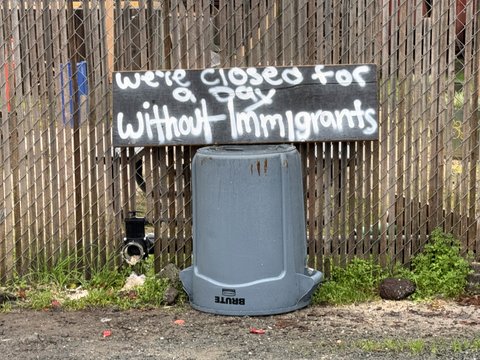 a wooden board with we'e closed for a day without immigrants spray painted on it. the sign is perched on an overturned gray trash can in front of a fence