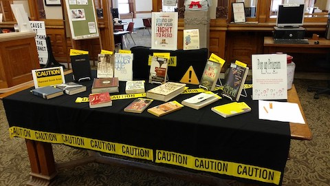 books on a table with yellow caution tape and signs that read caution banned book area and stand up for your right to read. some of the books are to kill a mockingbird by harper lee, the kite runner by khaled hosseini, beloved by toni morrison, the absolutely true diary of a part time indian by sherman alexie and the bible