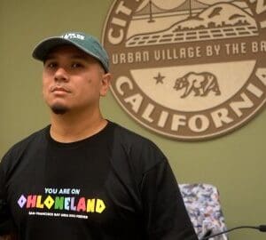 Robin López: How Albany’s First Latino and Indigenous Mayor Is Shaping the Future of His City a latino and indigenous man wearing a black T shirt that says "you are on ohlone land." Behind him is the seal of albany california which says it is an "urban village by the bay"