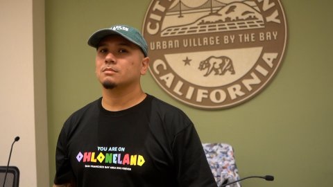 a latino and indigenous man wearing a black T shirt that says "you are on ohlone land." Behind him is the seal of albany california which says it is an "urban village by the bay"