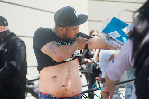 a latino man with his shirt pulled up revealing multiple scars on his torso and pointing to a scar on his upper arm