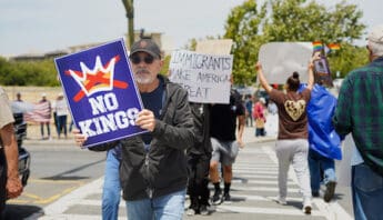 Protesters Rally in Hercules for ‘No Kings Day’ Demonstration white man with gray beard and mustache carrying "no kings" sign through crosswalk. behind him another person carries a sign that reads immigrants make america great. other people, signs, a US flag and rainbow flags are visible