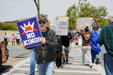 white man with gray beard and mustache carrying "no kings" sign through crosswalk. behind him another person carries a sign that reads immigrants make america great. other people, signs, a US flag and rainbow flags are visible