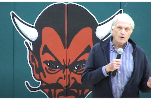 an older white man holding a microphone in front of an illustration of the devil