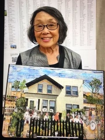 an older japanese woman holding an artwork she created with an illustration of a house with several people and a picket fence in front of it