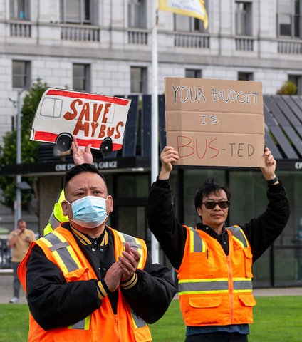 Two Asian men in orange safety vests. one is holding a cardboard sign that reads your budget is bus-ted. behind the other someone holds a gray and red sign made to look like a Muni bus with a message that reads save my bus