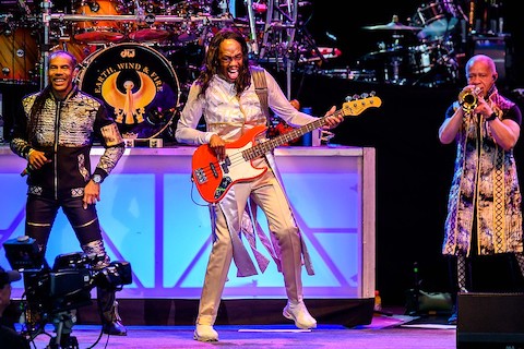 three black male musicians on stage, one with a microphone, one with a red and white bass guitar who appears to be screaming, and one playing a horn. a drum kit behind them says earth, wind & fire