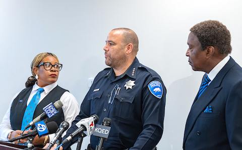 a black woman, a white man in a police uniform at a lectern with microphones for multiple san francisco bay area stations, and a black man in a suit
